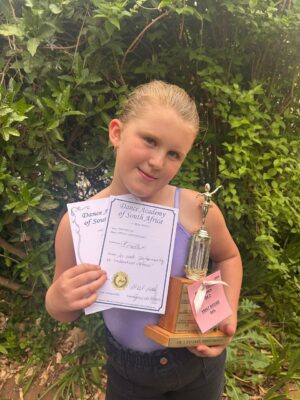 A young girl smiling and holding a dance trophy and certificates outdoors. She wears a purple top, standing against a background of lush green plants.