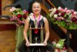 A young dancer in a black tutu holds a large trophy, smiling proudly. Behind her are bouquets of colorful flowers, creating a celebratory atmosphere.