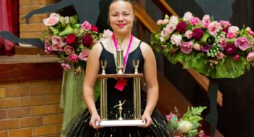 A young dancer in a black tutu holds a large trophy, smiling proudly. Behind her are bouquets of colorful flowers, creating a celebratory atmosphere.