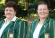 Two women smile at the camera, wearing green blazers with yellow stripes and a crest. They're outdoors, with lush greenery in the background.