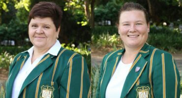 Two women smile at the camera, wearing green blazers with yellow stripes and a crest. They're outdoors, with lush greenery in the background.
