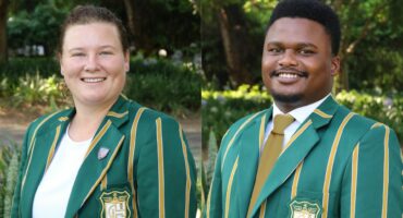 Two people wearing green and yellow-striped blazers with a crest, smiling outdoors against a backdrop of greenery, conveying a positive tone.