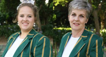 Two women standing outdoors, wearing green blazers with yellow stripes. The background features lush greenery, conveying a serene, natural setting.