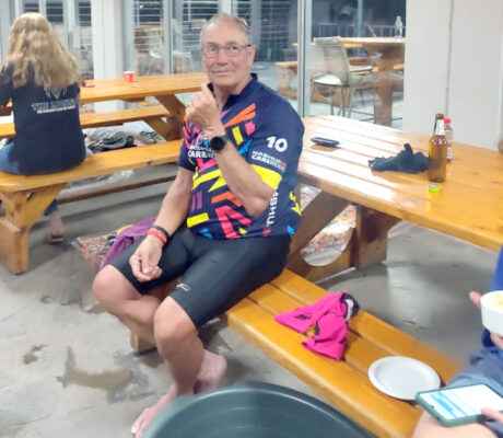 A man in colorful cycling gear sits on a wooden picnic bench, smiling and giving a thumbs-up. Casual setting with relaxed, friendly vibes.
