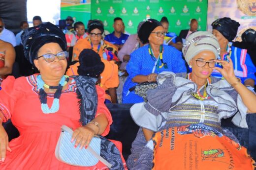 Two women in vibrant traditional attire and headwraps sit in a colorful crowd at an indoor event. They appear engaged and content.