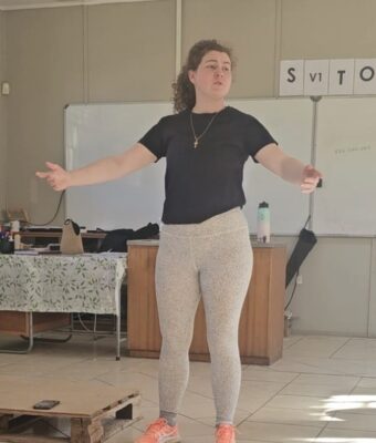 Woman in a black shirt and light gray leggings stands with arms outstretched in a classroom. A cross necklace and orange sneakers add color.