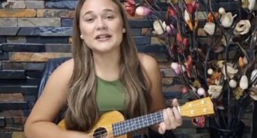 A person with long brown hair plays a ukulele while smiling, seated in front of a stone wall and decorative branches with colorful flowers. The mood is cheerful.