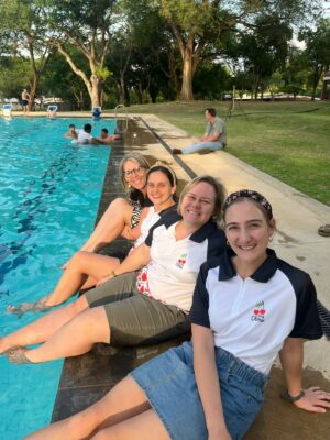 Four women sit smiling by a pool, dipping their feet in the water on a sunny day. They're wearing matching shirts, with trees and grass in the background.