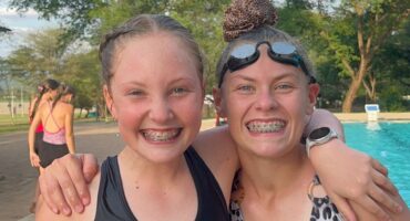 Two smiling girls with braces stand arm-in-arm at a poolside, wearing swimsuits. Trees and a bright sky are in the background, conveying a joyful summer vibe.