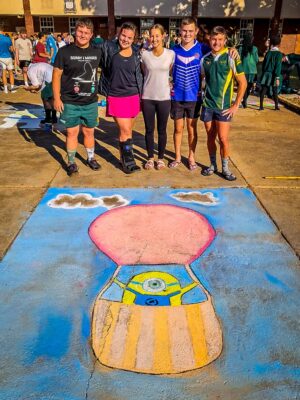 Five people smile, standing behind a colorful sidewalk chalk drawing of a hot air balloon with a Minion inside. The scene conveys joy and creativity.