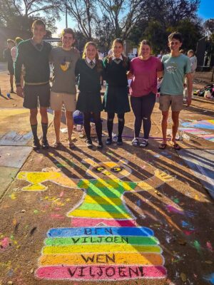 A group of six young people stands smiling on a colorful, chalk-drawn trophy on pavement. The art is vibrant and lively, expressing joy and camaraderie.
