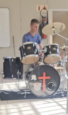 A young man in a blue shirt plays a black drum set with cymbals. The bass drum has a red cross and graphics. Sunlight filters into the room.