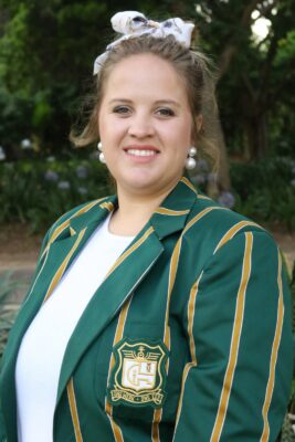 Smiling woman outdoors wearing a green blazer with gold stripes and an emblem. The background is lush with green foliage, conveying a cheerful tone.