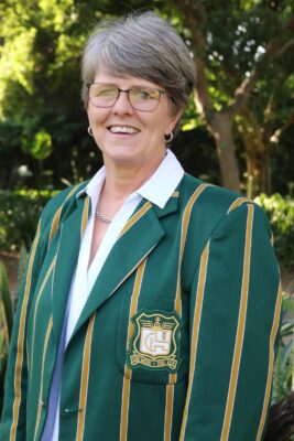 A woman with short gray hair, wearing glasses, smiles while standing outdoors. She is dressed in a green blazer with gold stripes, featuring a crest. The background is lush greenery, suggesting a park setting.
