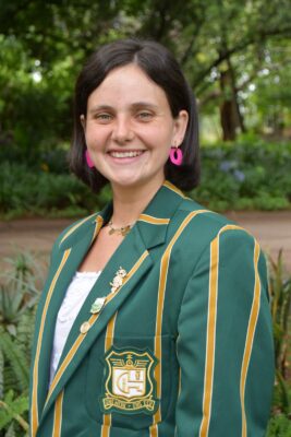 Smiling woman outdoors wearing a green blazer with gold stripes and an emblem. The background is lush with green foliage, conveying a cheerful tone.