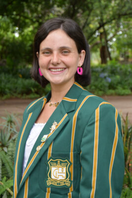 Young woman smiling outdoors, wearing a green blazer with gold stripes and school emblem. Pink earrings add a pop of color. Lush greenery in background.