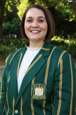 Smiling woman outdoors wearing a green blazer with gold stripes and an emblem. The background is lush with green foliage, conveying a cheerful tone.