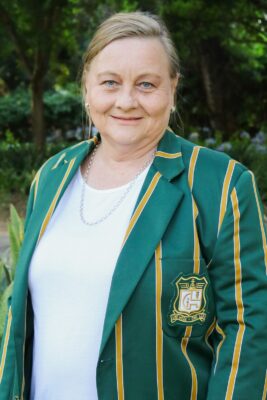 Smiling woman outdoors wearing a green blazer with gold stripes and an emblem. The background is lush with green foliage, conveying a cheerful tone.