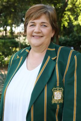 Smiling woman outdoors wearing a green blazer with gold stripes and an emblem. The background is lush with green foliage, conveying a cheerful tone.