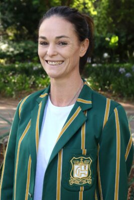 Smiling woman outdoors wearing a green blazer with gold stripes and an emblem. The background is lush with green foliage, conveying a cheerful tone.
