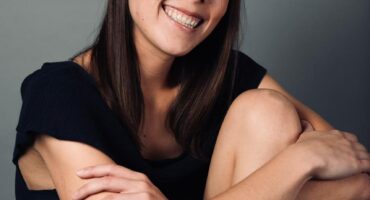 Young woman with long brown hair smiles warmly at the camera, seated with arms wrapped around her knees. She wears a dark top and light shorts against a neutral background.