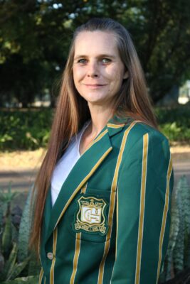 Smiling woman outdoors wearing a green blazer with gold stripes and an emblem. The background is lush with green foliage, conveying a cheerful tone.