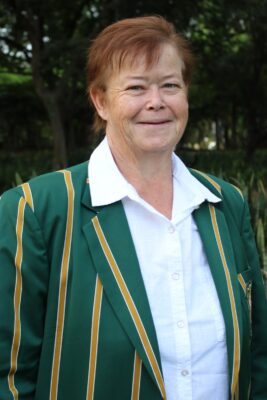 A person with short reddish hair smiles softly, wearing a green blazer with gold stripes and a white shirt, standing in a lush outdoor setting.