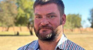 A man with short hair and a beard stands outside, wearing a checked shirt. He looks at the camera with a slight smile. Blurred trees and grass are in the background under a clear blue sky, conveying a calm, warm atmosphere.
