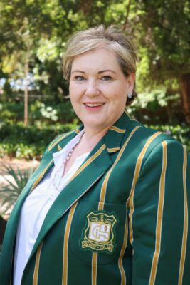 Smiling woman outdoors wearing a green blazer with gold stripes and an emblem. The background is lush with green foliage, conveying a cheerful tone.