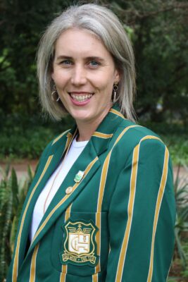 Smiling woman outdoors wearing a green blazer with gold stripes and an emblem. The background is lush with green foliage, conveying a cheerful tone.