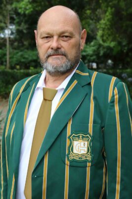 A man with a bald head and beard wearing a dark green jacket with yellow stripes and a crest, stands outdoors. He looks serious and confident.