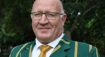 A smiling man in glasses wears a green blazer with yellow stripes and a gold tie, standing outdoors with greenery in the background.