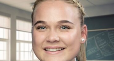 Smiling student in a green school uniform with badges, braided hair, standing in a classroom. Bright, cheerful atmosphere with windows and a chalkboard.