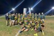 A rugby team poses on a field at night, wearing green and yellow jerseys, with coaches in jackets. Bright lights illuminate the scene, conveying joy.