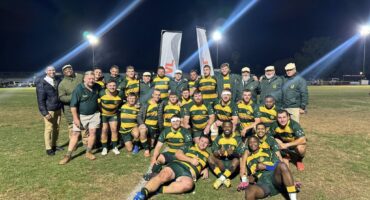 A rugby team poses on a field at night, wearing green and yellow jerseys, with coaches in jackets. Bright lights illuminate the scene, conveying joy.