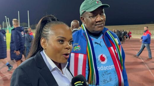 A woman in a suit energetically speaks into a microphone on a sports field, while a man in a colorful sports jacket and cap listens, surrounded by others.