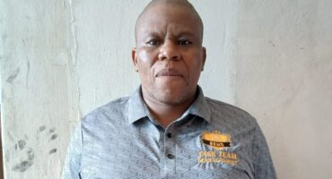 A person in a gray patterned polo shirt stands against a plain, textured wall. The expression is neutral, conveying a calm and composed tone.