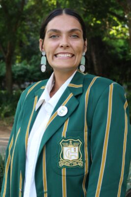 Smiling woman outdoors wearing a green blazer with gold stripes and an emblem. The background is lush with green foliage, conveying a cheerful tone.