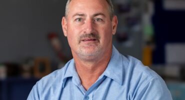 A middle-aged man with short hair and a mustache is wearing a light blue shirt with "CVO Marmer Personeel" embroidered. The background is blurred.
