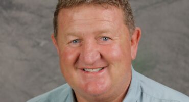 Smiling man in a gray shirt with short brown hair against a gray textured background. The mood is friendly and approachable.
