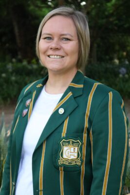 Smiling woman outdoors wearing a green blazer with gold stripes and an emblem. The background is lush with green foliage, conveying a cheerful tone.