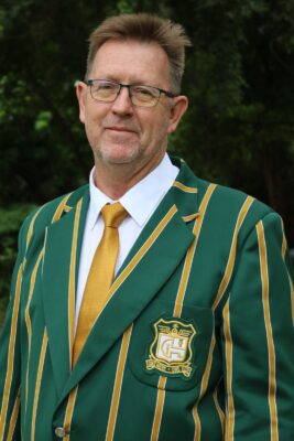 Man in glasses wearing a green blazer with yellow stripes and a matching tie. A badge on the blazer reads "GH." Background shows dense greenery.