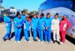 A group of people in blue tracksuits and casual attire pose cheerfully beside a tour bus on a sunny day, conveying a vibrant and positive energy.