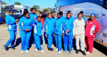 A group of people in blue tracksuits and casual attire pose cheerfully beside a tour bus on a sunny day, conveying a vibrant and positive energy.