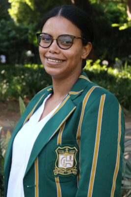 Smiling woman outdoors wearing a green blazer with gold stripes and an emblem. The background is lush with green foliage, conveying a cheerful tone.