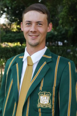 A man in a green blazer with yellow stripes and a matching tie smiles warmly. The blazer features a school emblem. The background is lush greenery.