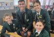 Four smiling students in green school uniforms gather around a table with a laptop. They have badges on their blazers, creating a cheerful classroom scene.