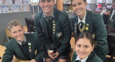 Four smiling students in green school uniforms gather around a table with a laptop. They have badges on their blazers, creating a cheerful classroom scene.