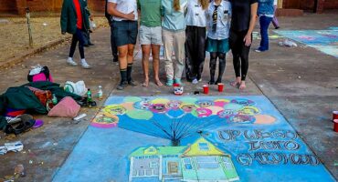 A group of six smiling people stands barefoot on the pavement, showcasing bright chalk art inspired by the movie "Up" with colorful balloons and a house. The art reads "Up We Go Into Childhood," conveying a playful and nostalgic tone.