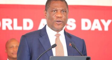 A man in a suit and red ribbon pin speaks at a podium against a red backdrop reading "World AIDS Day." He looks serious and focused.